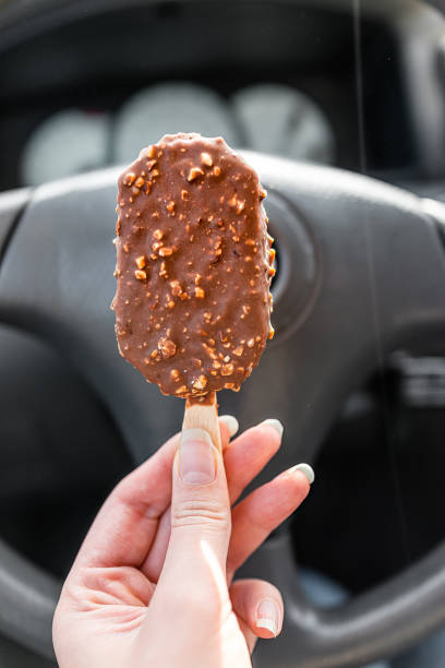 держа в руке один шоколадный миндальный ореховый батончик мороженого на палочке вертикального вида с фоном рулевого колеса в поездке - people snack almond brown стоковые фото и изображения