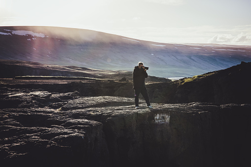 Mid adult Photographer standing close to a cliff of rural central iceland in moody sunset light. Taking pictures of the vast icelandic moody sunset landscape. Solo Traveller, Photographer Outdoors Travel Concept. North Central Iceland, Nordic Countries, Northern Europe.