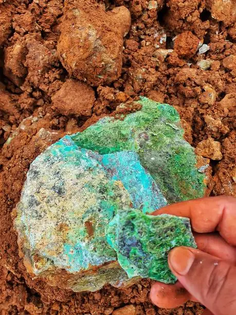 A man's hand is holding a green nickel rock on a brown background in a very hot and arid nickel mine