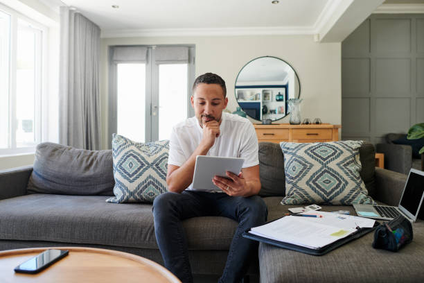 Shot of a young man sitting on his sofa at home and looking contemplative while using a digital tablet