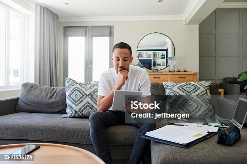 istock Shot of a young man sitting on his sofa at home and looking contemplative while using a digital tablet 1340636174