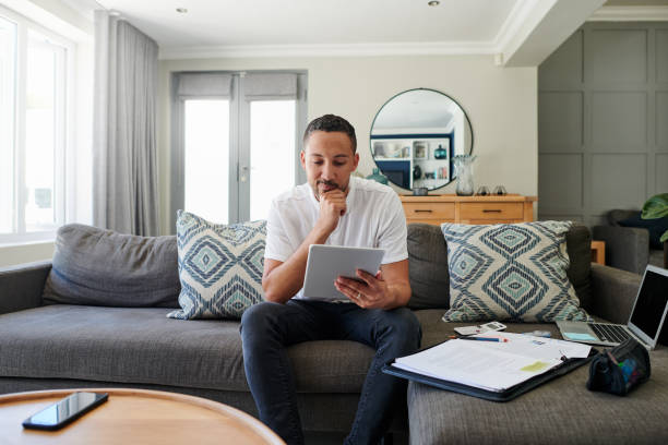 Shot of a young man sitting on his sofa at home and looking contemplative while using a digital tablet stock photo