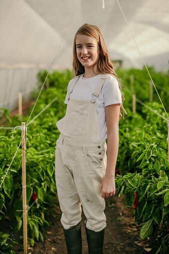 Portrait of a countryside tennage girl standing in the greenhouse while harvesting. Girl is wearing boots and a denim pants on suspenders.