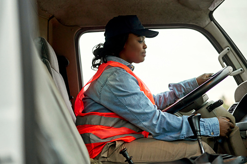 Side view of mid 20s Black woman in casual clothing, reflective vest, and cap sitting in driver’s seat of vehicle cab, ready to start road trip.