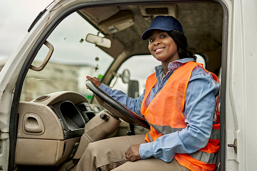 Three-quarter front view of Black woman in casual clothing, cap, and reflective vest sitting in driver’s seat of delivery truck and smiling at camera.