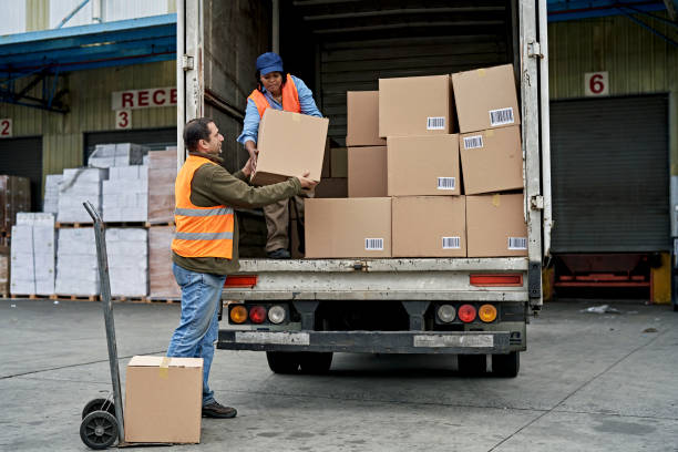 Black Female Truck Driver Loading Boxes in Cargo Space Full length view of late 40s man moving boxes from loading dock with hand truck and lifting into hands of woman organizing merchandise on vehicle. loading bay stock pictures, royalty-free photos & images