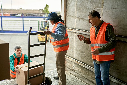 Mid adult man lifting cardboard box onto hand truck, senior man checking mobile device, and young woman preparing to organize merchandise within cargo space.