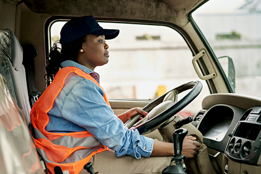 Side view of Black woman in casual clothing, cap, and reflective vest sitting in driver’s seat and preparing to begin road trip.