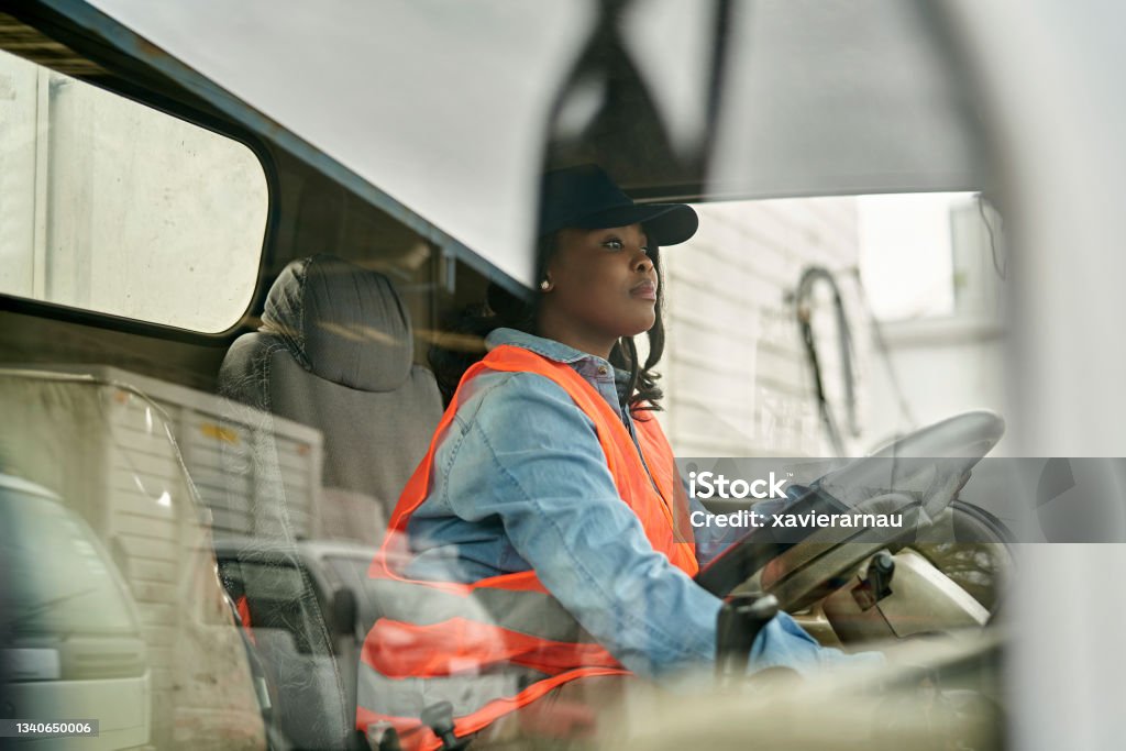 Black Female Truck Driver Photographed Through Window Personal perspective of mid 20s woman in casual clothing, cap, and reflective vest sitting in driver’s seat, looking straight ahead, ready to begin road trip. Truck Driver Stock Photo