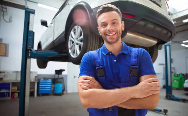 Confident handsome young and experienced car repair worker in work overalls posing against the background of lifted cars in a car service