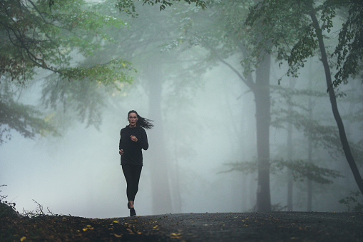 Young athletic woman running on the road in foggy forest. Copy space.