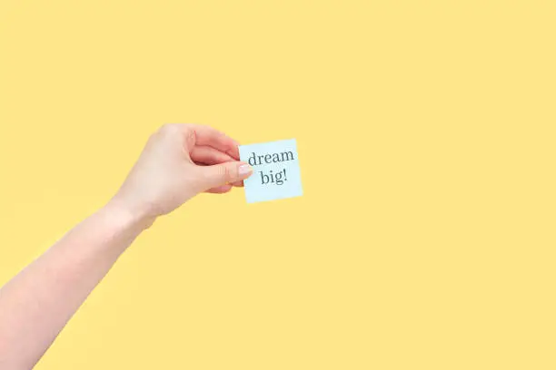 The hand holds a sticky note with inscription Dream big! Motivational statement.