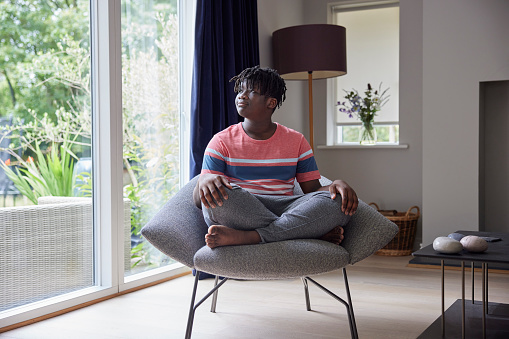 Thoughtful Teenage Boy Sitting Cross Legged On Chair At Home Looking Out Of Window