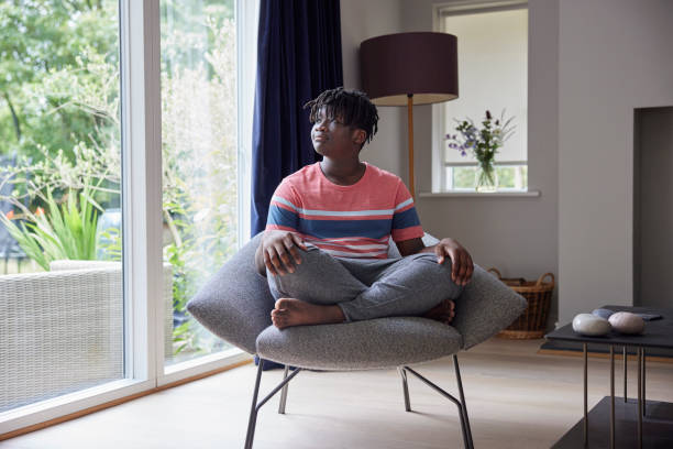 Thoughtful Teenage Boy Sitting Cross Legged On Chair At Home Looking Out Of Window Thoughtful Teenage Boy Sitting Cross Legged On Chair At Home Looking Out Of Window mindfulness children stock pictures, royalty-free photos & images