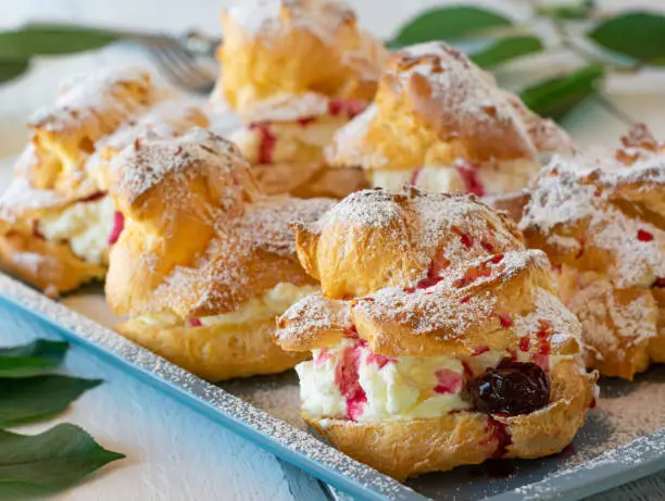 Homemade fresh baked cream puff pastry with whipped cream filling and sour cherries. Served on a tray on white wooden table. Closeup view with blurred background