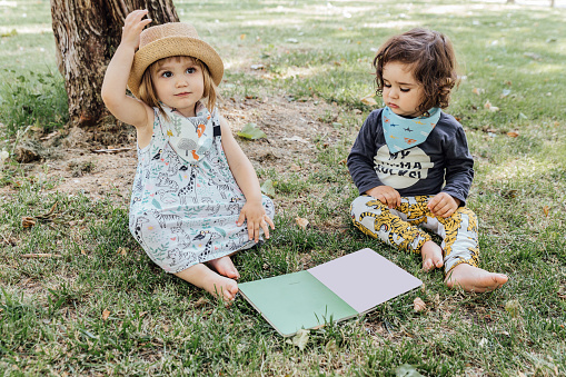 Little blonde girl in a hat looking up with her hand raised while her little friend looks down at a book in the park. Adorable little brother and sister sitting on grass in park and reading book together