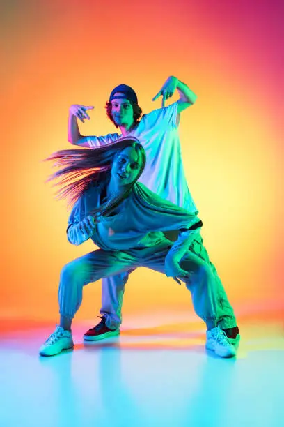 Photo of Young hip-hop dancers, stylish emotive girl and boy in action and motion in casual sports youth clothes on gradient multi colored background at dance hall in neon light.