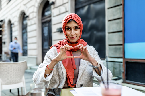 Beautiful business Muslim  woman drinks a cup of black coffee smiling in love, showing with her hands the symbol of the heart and the shape. Romantic concept.