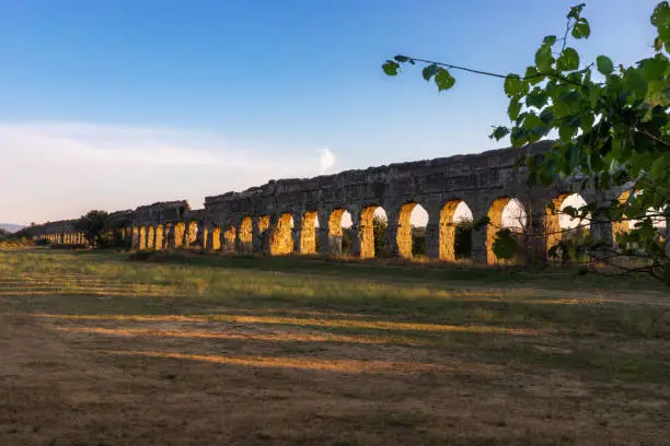 In the park of the ancient Roman aqueducts, the sun sets and the arches of the ancient Claudius aqueduct cast their shadow on the lawn in front.