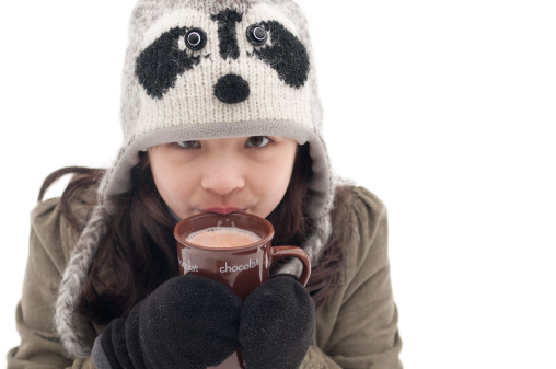A young girl wearing a knitted raccoon hat looks up at the camera while sipping a mug of hot chocolate on a snowy, winter day.  Melting snow can be seen on her hat and mittens.