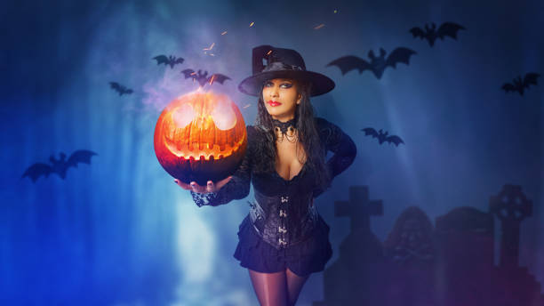 Halloween witch girl with magic pumpkin of spells portrait. Beautiful young woman in witches hat conjuring, making witchcraft. Over spooky dark magic forest background. Wide Halloween party art design. stock photo