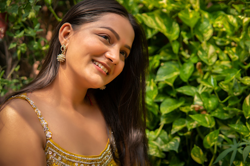 Outdoor portrait of beautiful, happy Indian young woman with shining log hair contemplating and looking away with a toothy smile. She is enjoying sunshine, fresh air in lush green nature at day time.