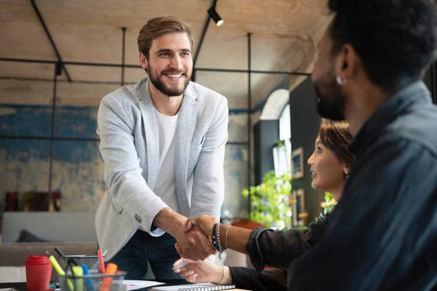 istock Two men shaking hands and looking at each other with smile while their coworkers sitting at the business meeting 1340692190
