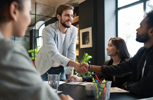 istock Two men shaking hands and looking at each other with smile while their coworkers sitting at the business meeting 1340692219