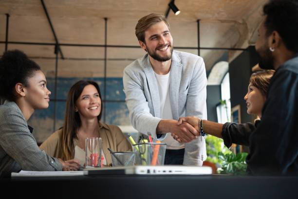 istock Two men shaking hands and looking at each other with smile while their coworkers sitting at the business meeting 1340692220