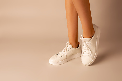 Casual footwear - white leather sneakers shoes and girlâs legs on nude background. Layout with free copy (text) space.