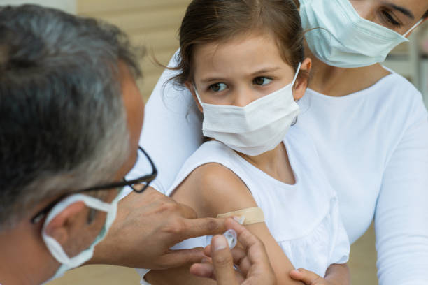 Vaccination Child with protective face mask is sitting in her mothers lap and  is getting vaccination from a doctor. hepatitis photos stock pictures, royalty-free photos & images