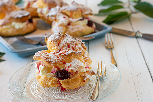Delicious homemade baked cream puff filled with whipped cream and sour cherries. Served on a plate with fork on white wooden table. Closeup view with blurred background.