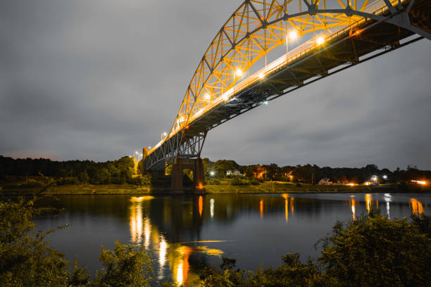 istock Gold-colored Sagamore Bridge over Cape Cod Canal in Massachusetts at dawn. Glowing bridge lights reflected on the tranquil water. Moody gray clouds passing over the arch. 1340710919