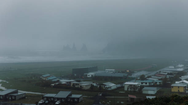 Dramatic view of the beach with rocky cliffs and the small town covered by summer fog in South Iceland