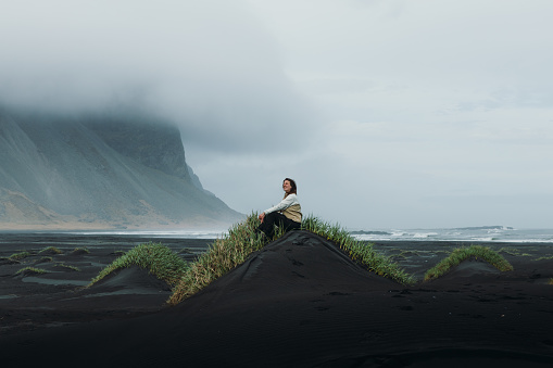 Silhouette of a young woman with long hair walking at the remote dramatic black beach with panoramic view of the mountains