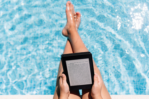 Top view of a relaxed girl reading an e-book on a digital tablet while sitting on the ledge of a swimming pool. Technology and summer concept.