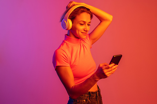 One caucasian girl in headphones putting hair up isolated over gradient background in neon lights. Music lifestyle. Concept of human emotions, facial expression, beauty. Copy space for ad