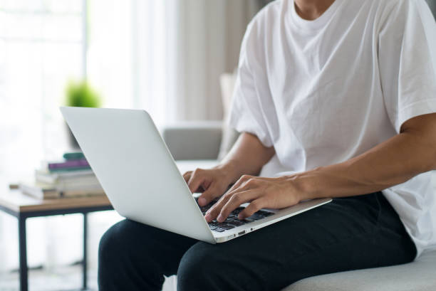 Young men wearing white t-shirt and black trousers sitting on sofa use laptops to work at home in living room. stock photo
