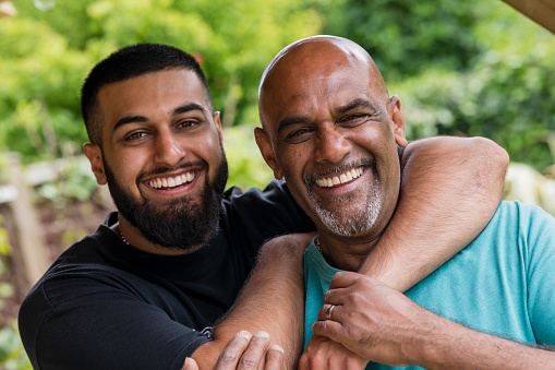 Pakistani father and son laughing with their arms around each other in the North East of England. They are relaxing in a garden in winter, looking at the camera.