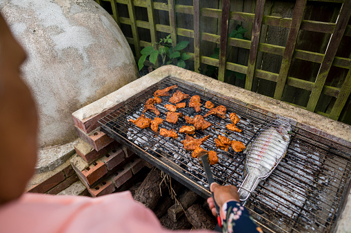 Over the shoulder view of a pakistani man in his garden in Middlesbrough, North East of England cooking on a BBQ. He is throwing a social gathering in summer. There are chicken wings and a fish cooking.