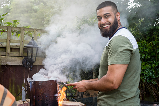 Pakistani man in his garden in Middlesbrough, North East of England cooking on a BBQ, looking at the camera. He is throwing a social gathering in summer.
