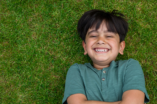 Close up of a young asian boy laying down on the ground on grass while playing in a domestic garden. He is in Middlesbrough in the North East of England.