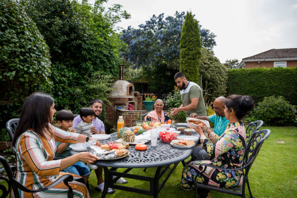 Family Eating in the Sun Together Asian/Pakistani multi generation family sitting around a table in a garden in Middlesbourgh, North East of England. There is a smart speaker on the table. britain british audio stock pictures, royalty-free photos & images