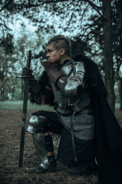 istock Medieval knight in armour standing on one knee in forest with his sword. 1340723330