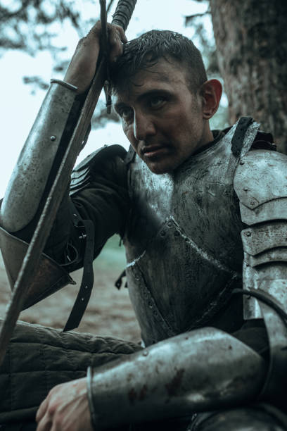 istock Medieval knight in armour sits and rests under tree with his sword. 1340723395