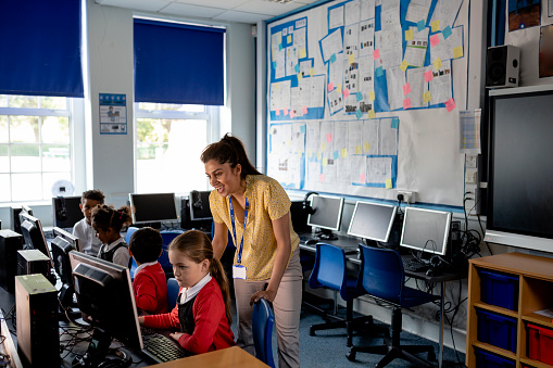 A wide-angle view of a female teacher and her young pupils working o the computers as part of their computing class in school. They are all waiting to be given instructions from the teacher.