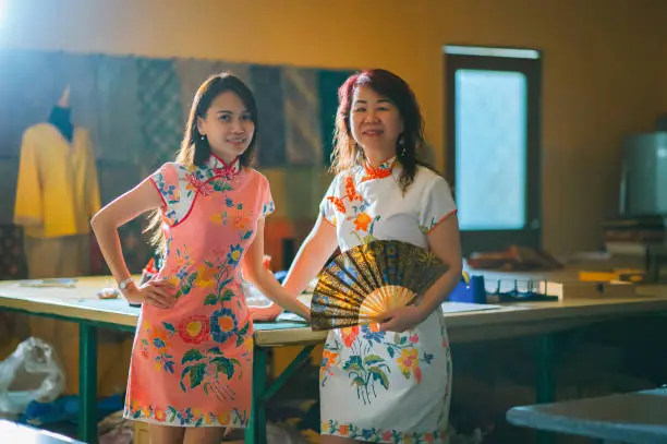 asian chinese female tailor small business owner and her mother standing at work place with chinese tradition clothing cheongsam holding folding fan hands on chin smiling with confidence and satisfaction looking at camera