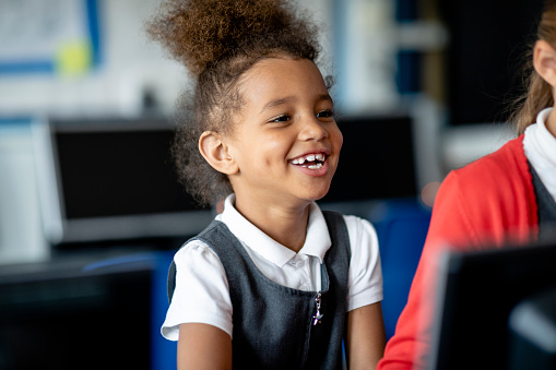 A close up of a little girl with curly brown hair with a big smile on her face as she gets to grips with the computer in class. She is wearing a school uniform and is sat at a desk next to her best friend in her class.