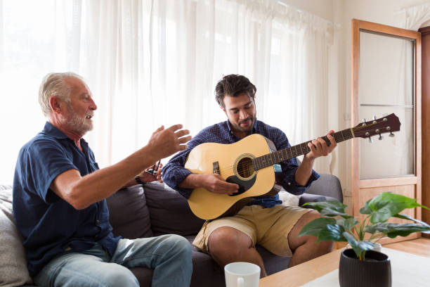 Happy Senior Father sing a song and Adult Son playing guitar on the sofa at home. Elderly father and son spending time together at home. Happy Senior Father sing a song and Adult Son playing guitar on the sofa at home. Elderly father and son spending time together at home. father and son guitar stock pictures, royalty-free photos & images