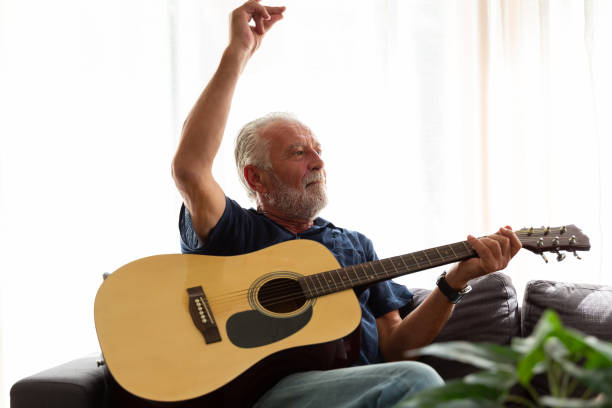 istock Cheerful senior man playing guitar while sitting on sofa at home. Elderly man learning guitar on holiday. people, quarantine, holiday concept 1340736840
